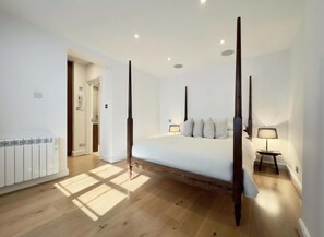 A bedroom with a four-poster bed, a radiator, a nightstand with a lamp, and a door leading to a bathroom. - Heritage South Ken with Fireplace, Super King Bed (London)