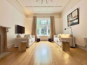 A spacious room with two white sofas, a fireplace, a large window with curtains, a floor lamp, and a painting on the wall. - Heritage South Ken with Fireplace, Super King Bed (London)