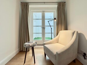 A room with a white armchair, a small table with a magazine, a floor lamp, and a door with curtains. - Heritage South Ken with Fireplace, Super King Bed (London)