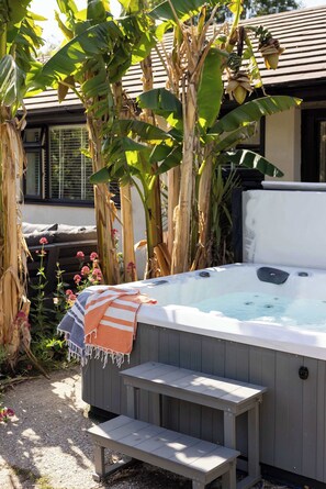 A hot tub with steps and a bench, surrounded by banana trees and flowering plants. - Scandi-Style Birdham Retreat with Hot Tub & Garden (Birdham)