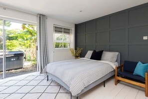 A bedroom with a bed, a sofa, a window with blinds, a radiator, and a view of greenery outside. - Scandi-Style Birdham Retreat with Hot Tub & Garden (Birdham)