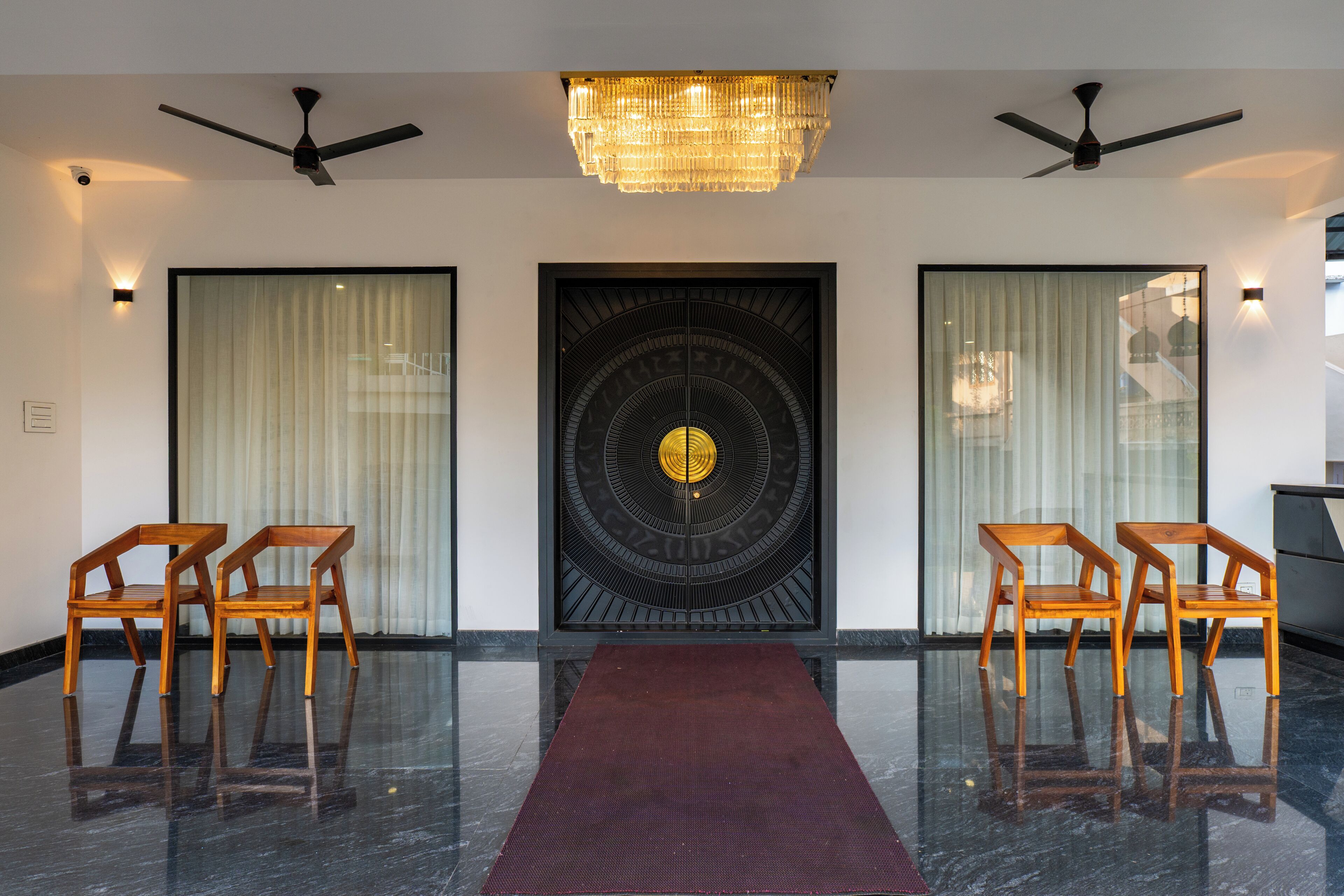 A lobby with a dark marble floor, a red carpet, two wooden chairs, a black door with a circular design, and ceiling fans.