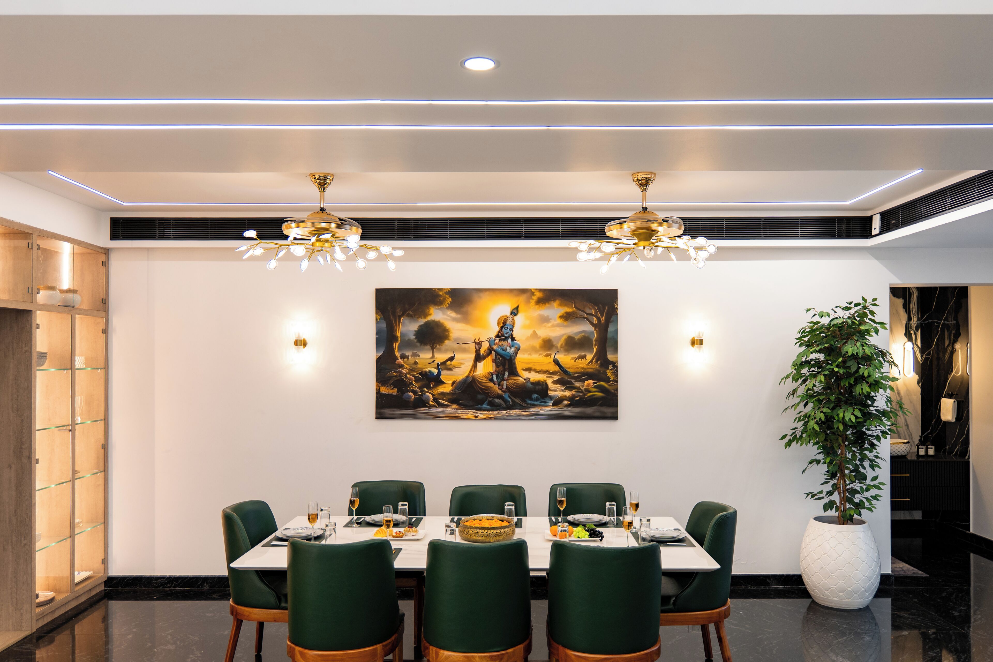 A dining area with a table set for six, green upholstered chairs, a large painting on the wall, and a potted plant.