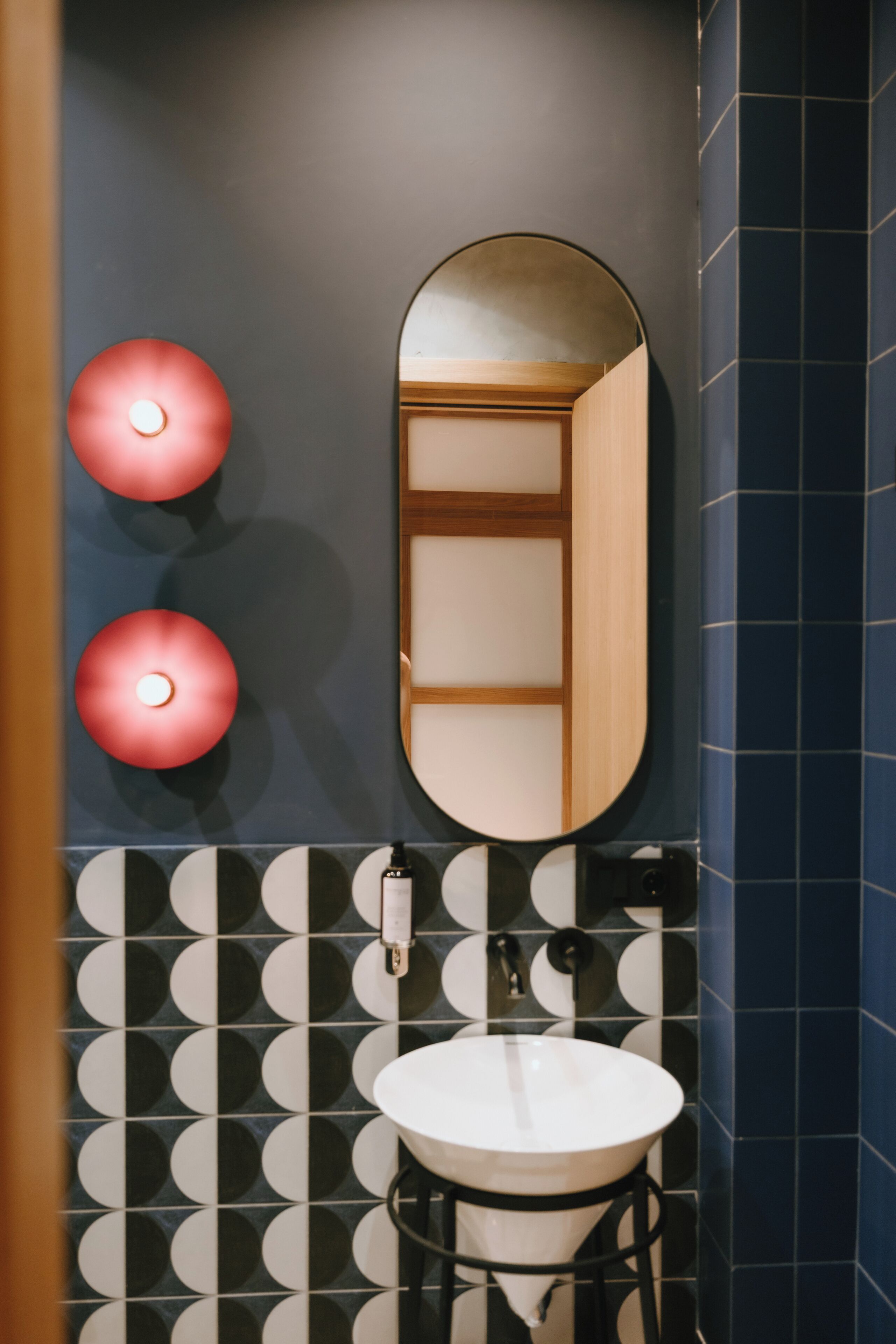 A bathroom with a round mirror, two red lights, a sink, and patterned tiles.