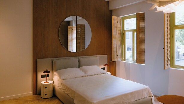 A bedroom with a bed, a bedside lamp, a nightstand, a mirror, and a window with shutters.