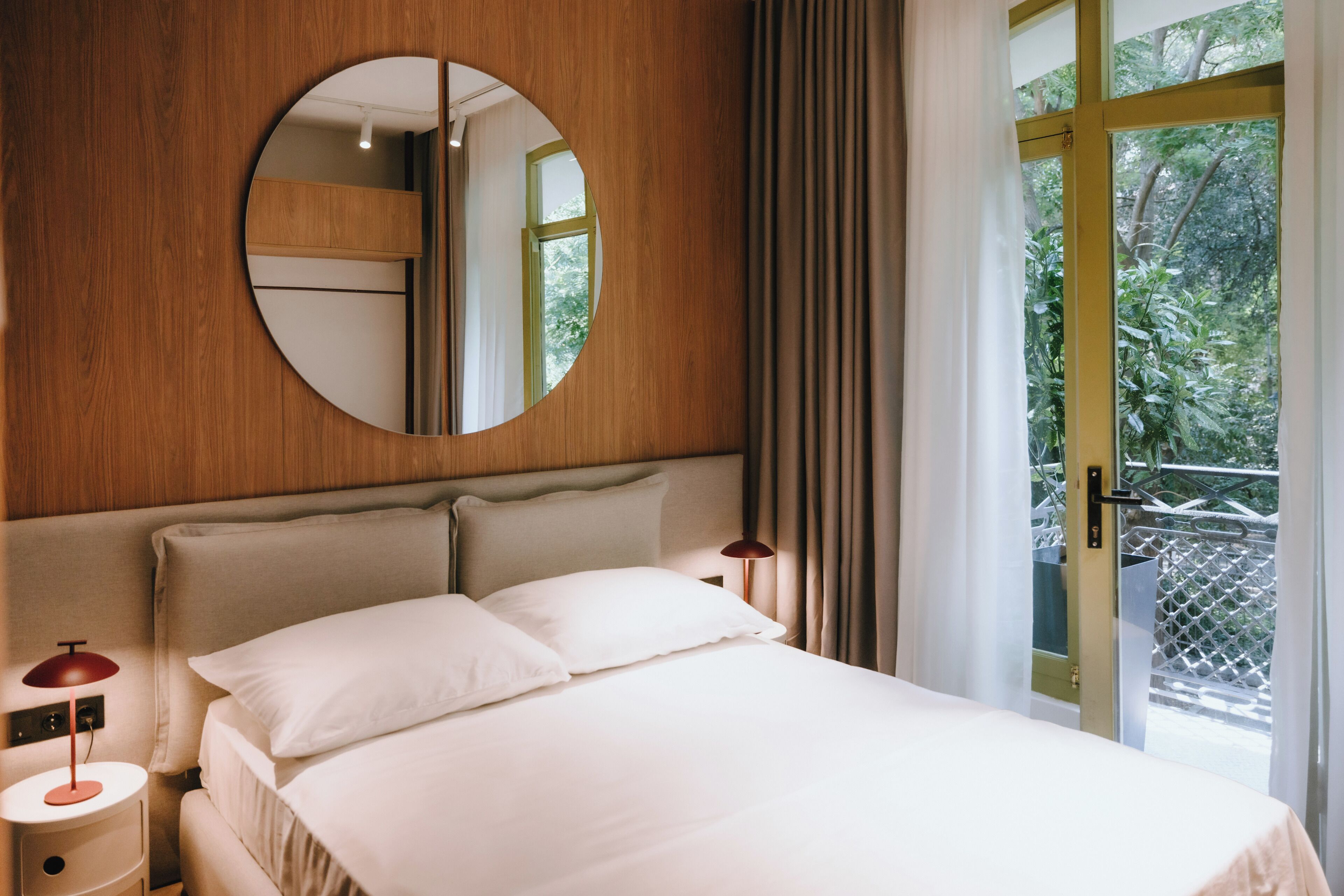 A hotel room with a bed, a round mirror, a nightstand, a lamp, and a view of greenery through a window.