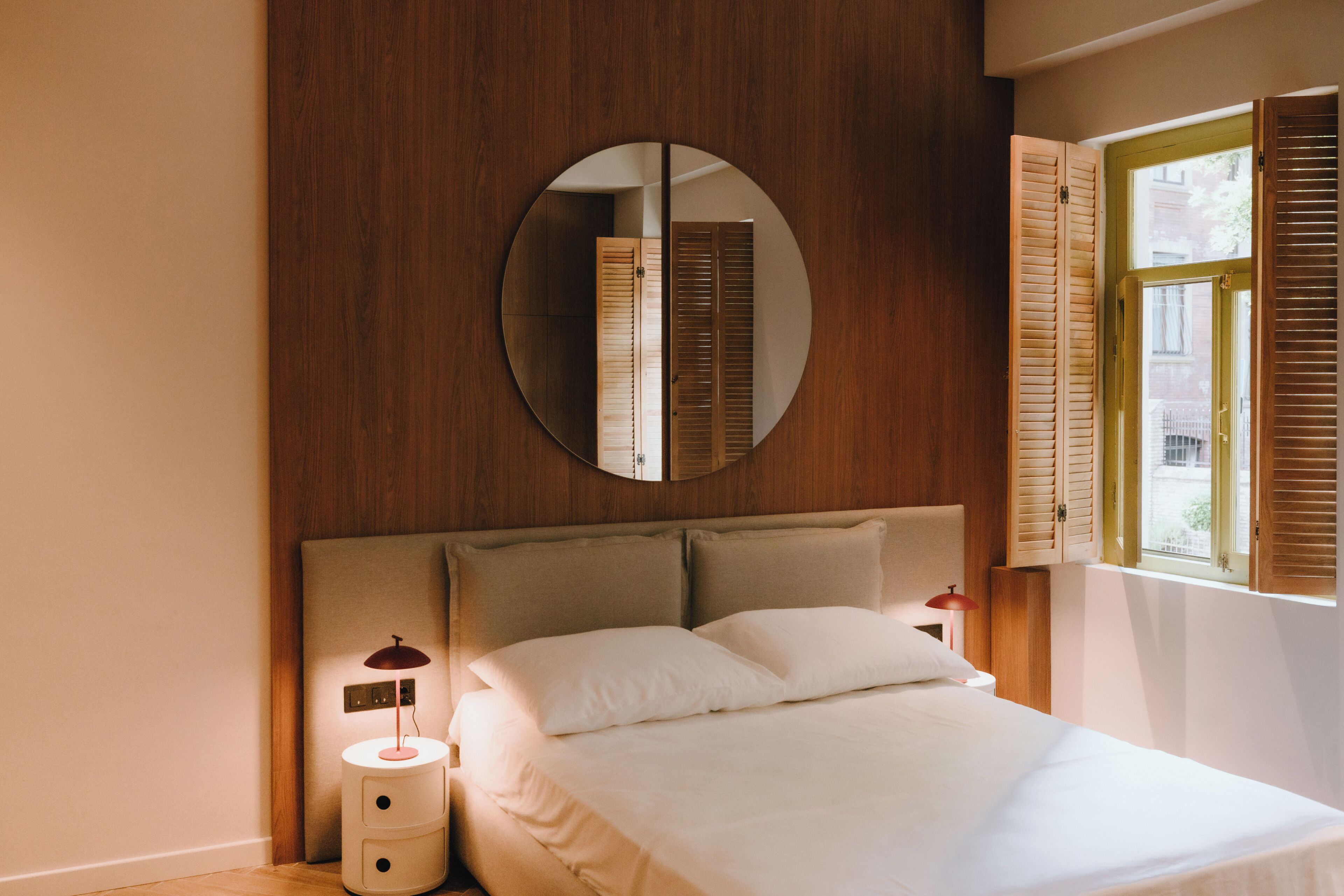 Une chambre à coucher avec un lit, une table de chevet avec une lampe, un miroir et une fenêtre avec des volets.