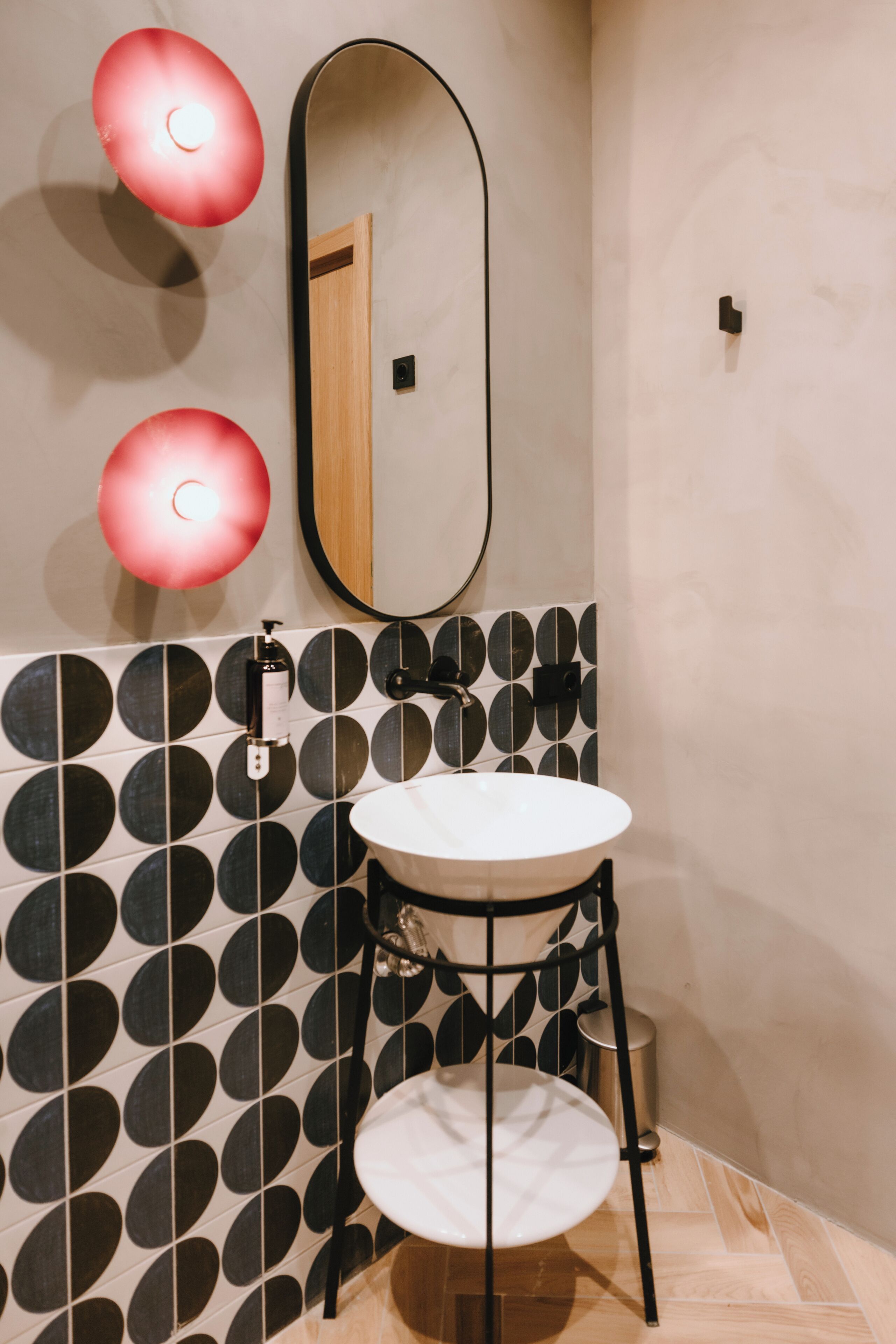 A bathroom with a white sink, black circular wall tiles, a wooden door, and two red wall-mounted lights.