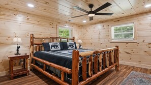 Quarto de cabana de madeira com uma cama, uma mesinha de cabeceira, um abajur, um ventilador de teto e uma janela com persianas.