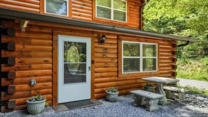 Eine Blockhütte mit einer Haustür, zwei Fenstern, einer kleinen Veranda mit einer Bank und Kübelpflanzen.