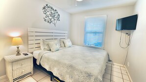 Un dormitorio con una cama, una mesita de noche, un televisor montado en la pared y una ventana con persianas.