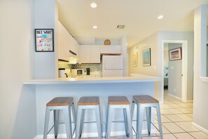 A modern kitchen with a central island, bar stools, and a clock on the wall. - Welcome to Shipyard Unit 155, a charming 2-bedroom, 1-bath ground-level condo located in the desirable Truman Annex community, just two blocks from Duval Street. This convenient Key West is within easy walking distance of the Truman Waterfront. (Key West)