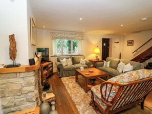 A cozy living room with a stone fireplace, a wooden coffee table, a floral-patterned armchair, and a sofa with cushions. - The Chimes (Hope Valley)