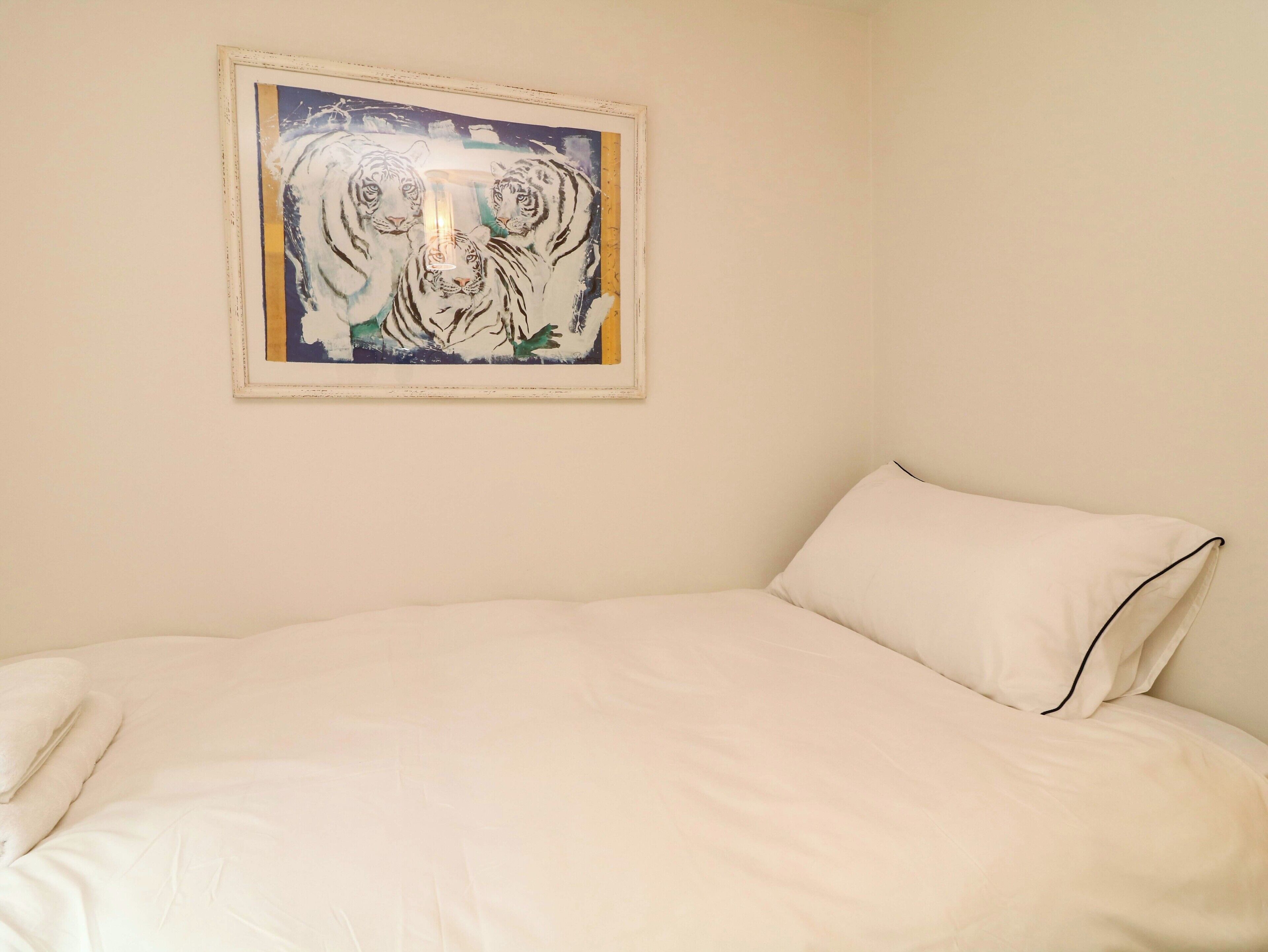 A bedroom with a bed, a framed painting of tigers, and a plain wall.