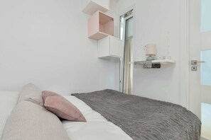 Een slaapkamer met een bed, kussens, een grijze deken, een witte deur en een klein plankje met decoratieve objecten.