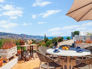 Eine Außenpatio mit einem runden Tisch, Korbstühlen, Topfpflanzen und einem schönen Blick auf Meer und Berge.