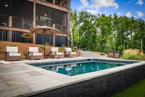 Ein Schwimmbecken mit Steinrand, umgeben von einer Terrasse mit Stühlen und einem Tisch.
