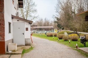 A gravel path leading to a white building with a door, surrounded by greenery and a fenced area with a wooden structure in the background.