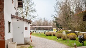 En grussti der fører til en hvid bygning med en dør, omgivet af grøn vegetation og et indhegnet område med en trækonstruktion i baggrunden.
