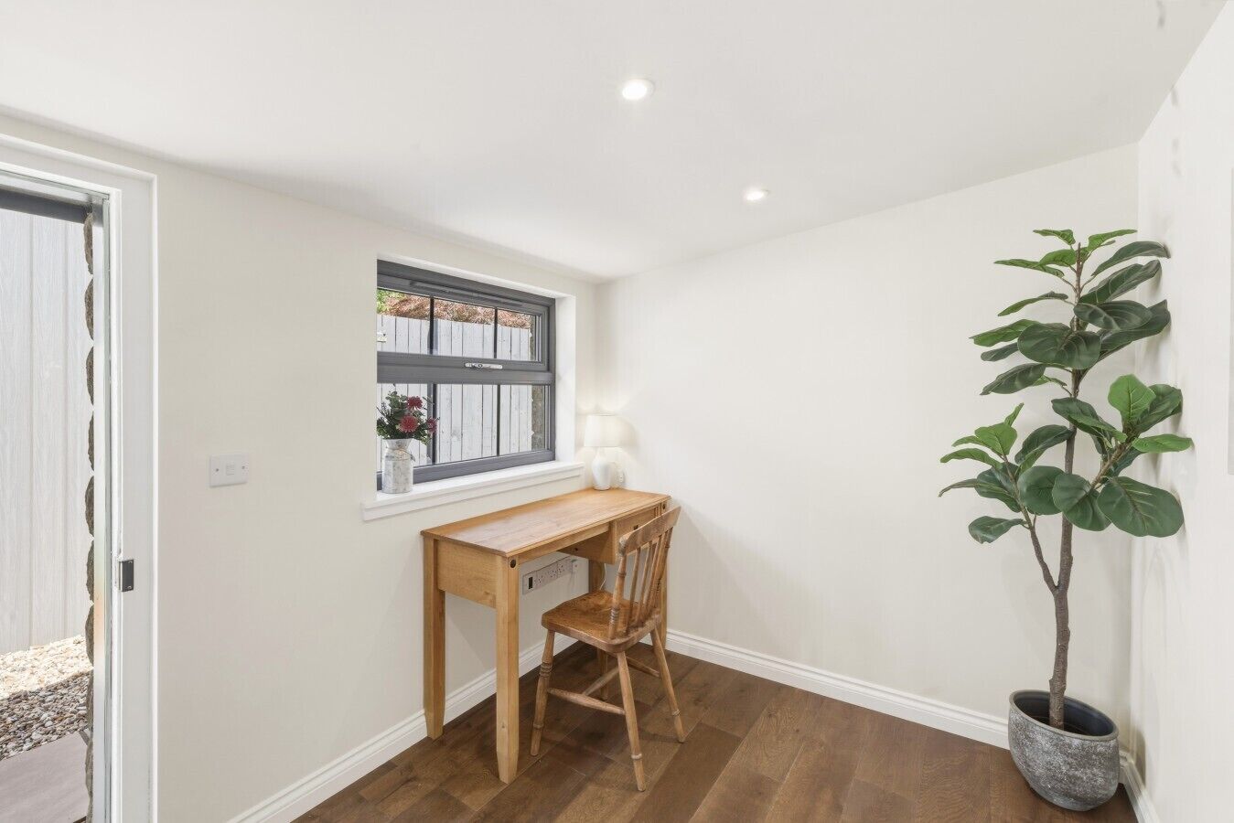 A room with a wooden desk, a chair, a potted plant, and a window with a view of a fence and flowers.