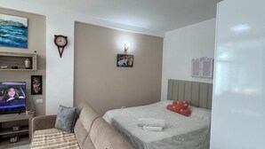 Ein modernes Wohnzimmer mit einer Couch, einem Flachbildfernseher, einem Bett mit weißer Bettdecke und einer Wanduhr.