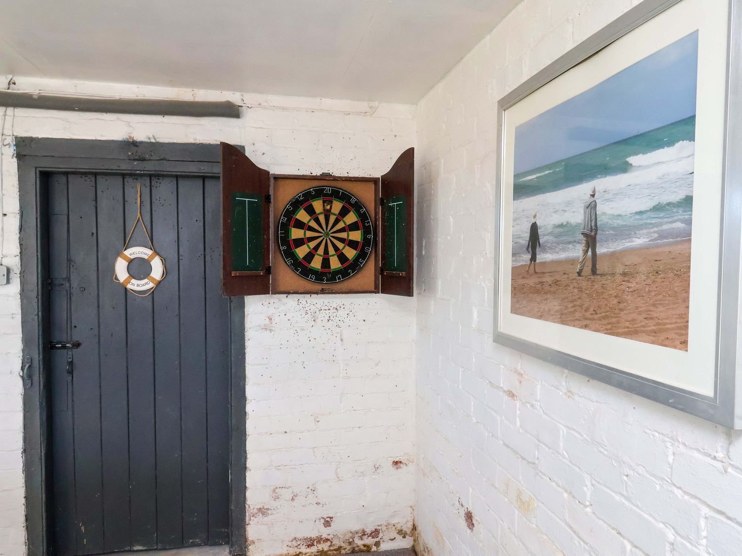 A room with a dartboard, a door, and a framed picture of a beach scene.