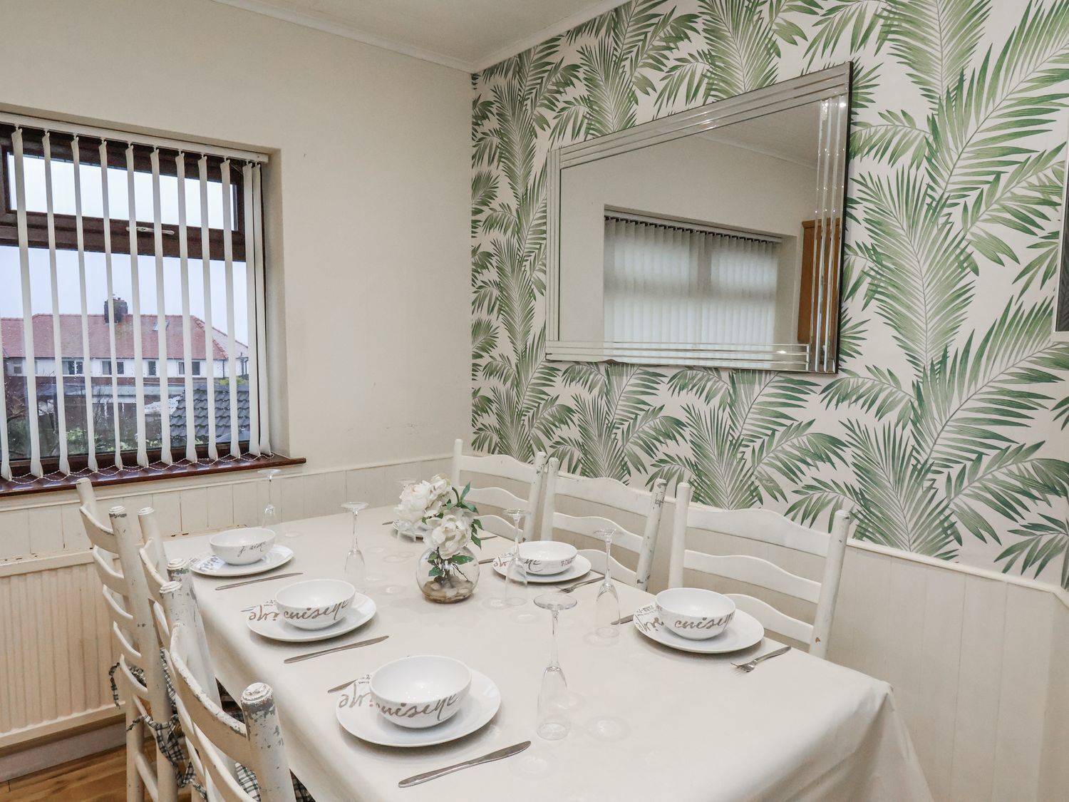 A dining room with a table set for six, white chairs, a window with blinds, and a wall with a leaf pattern.