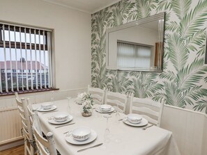 A dining room with a table set for six, white chairs, a window with blinds, and a wall with a leaf pattern. - Beachcrest (Whitby)