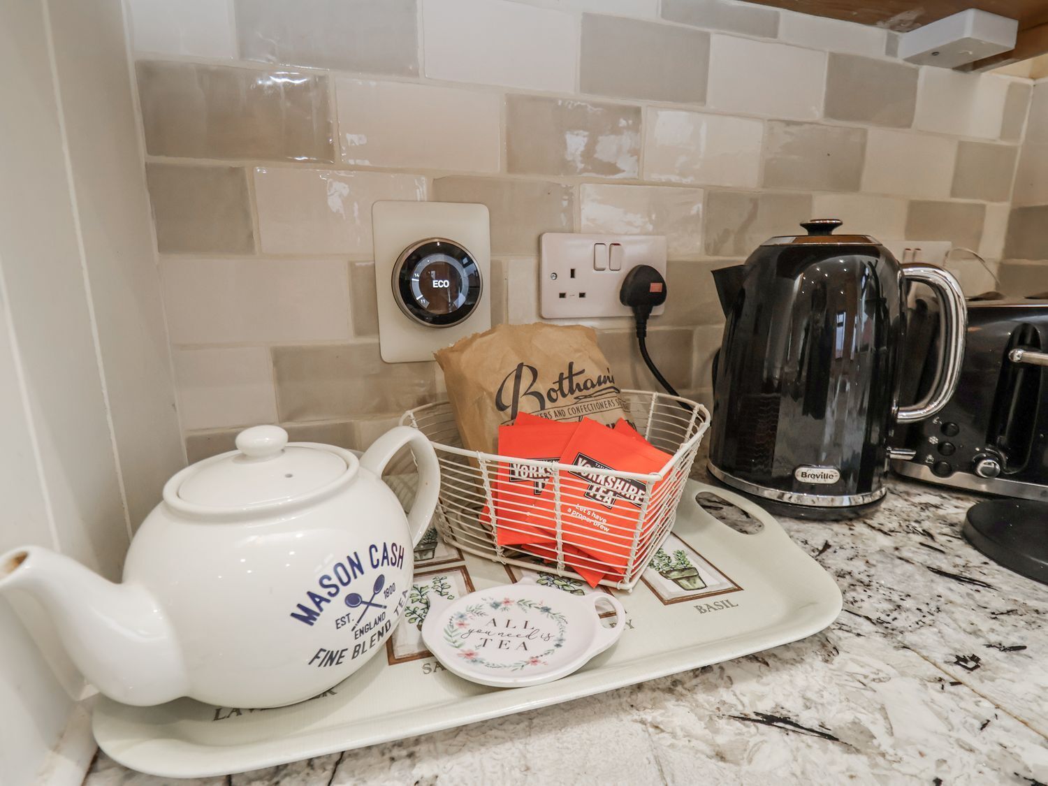A kitchen counter with a white teapot labeled 'Mason Cash,' a basket of red tea bags, a coffee maker, and a kettle.
