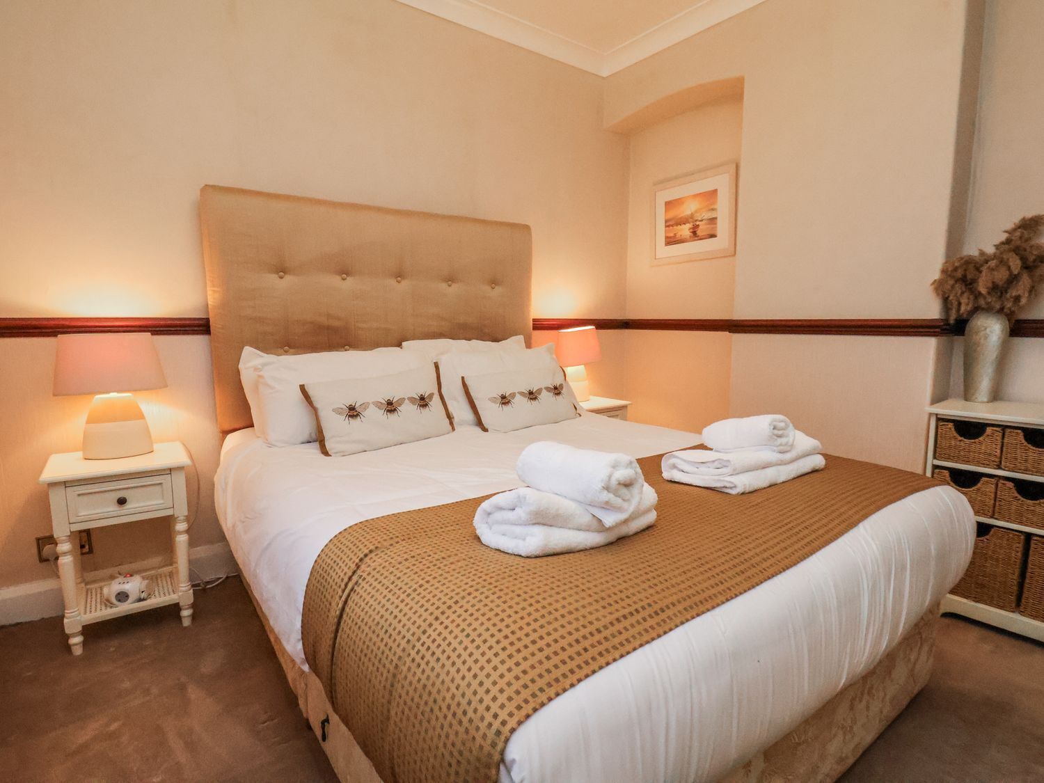 A neatly made bed with a brown headboard, white bedding, and a decorative pillow with a bee design. There are two bedside tables with lamps, a small drawer, and a vase with dried flowers on a nightstand.