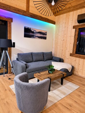 A cozy living room with a grey sofa, a wooden coffee table, and a striped armchair. A tripod lamp is visible to the left, and a framed picture hangs on the wall. - "LSN6" Chalet: Cozy & High-End Comfort with Sauna, Spa, Air Conditioning, Foosball, and Pool (Anould)