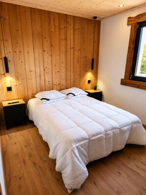 A bedroom with a wooden wall panel, a bed with white bedding, two bedside tables with lamps, and a window with a view. - "LSN6" Chalet: Cozy & High-End Comfort with Sauna, Spa, Air Conditioning, Foosball, and Pool (Anould)