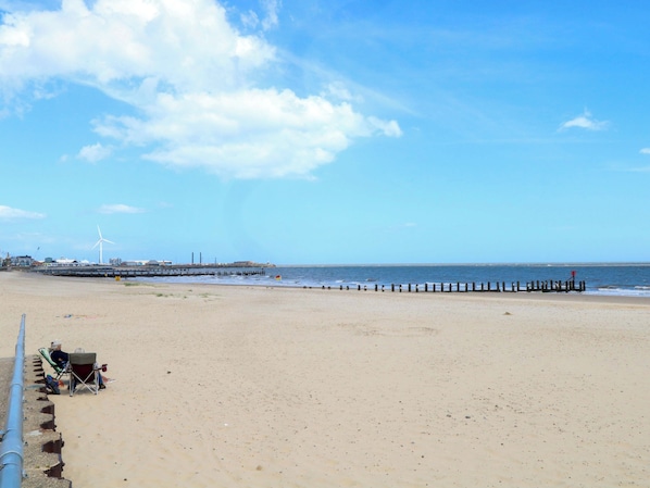 A wide sandy beach with a jetty extending into the water, a wind turbine in the background, and a clear blue sky.