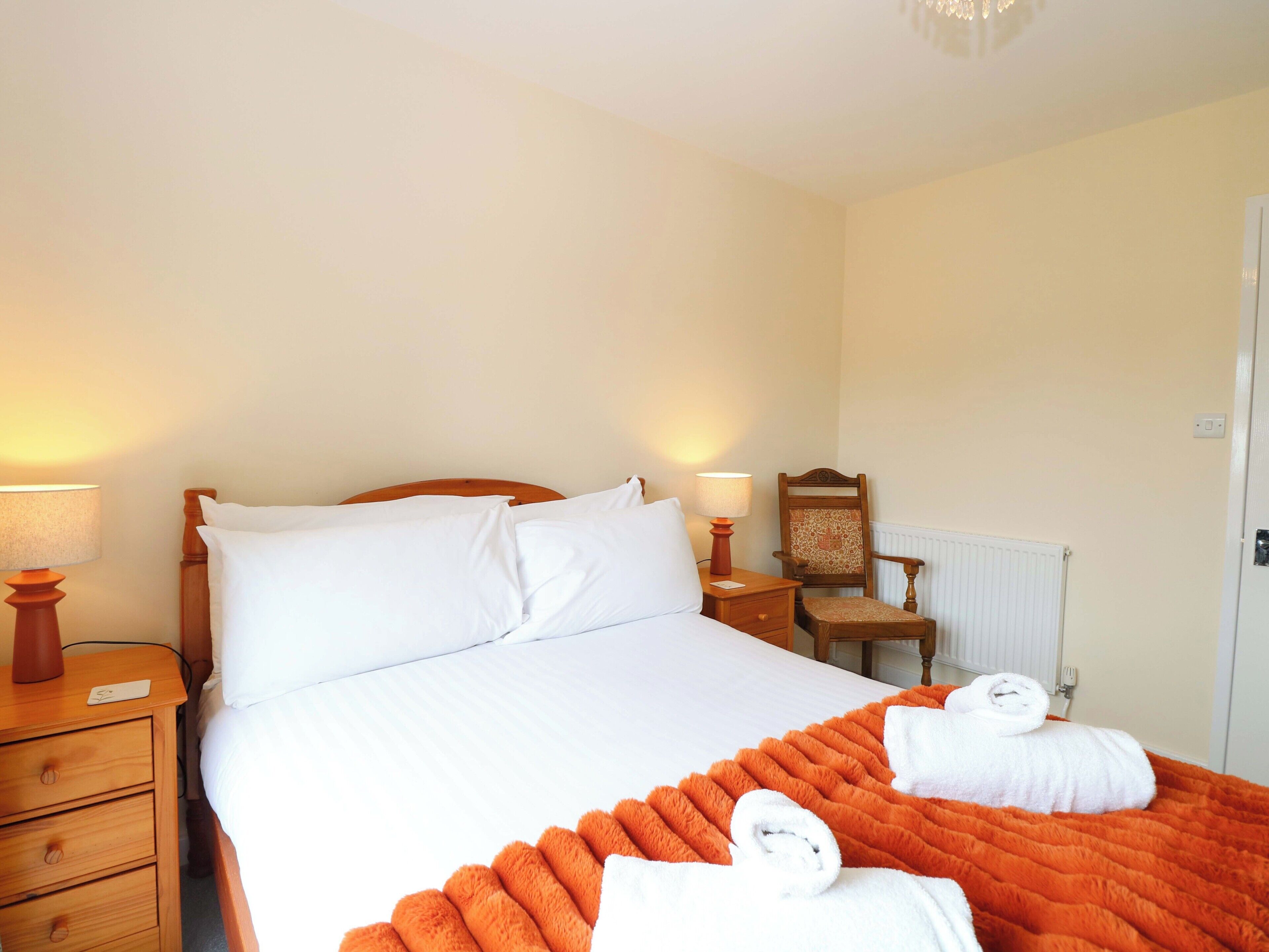 Una cama bien hecha con sábanas blancas, un edredón naranja y dos toallas. Hay dos mesitas de noche con lámparas, una silla de madera y un radiador.