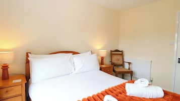 Una cama bien hecha con sábanas blancas, un edredón naranja y dos toallas. Hay dos mesitas de noche con lámparas, una silla de madera y un radiador.