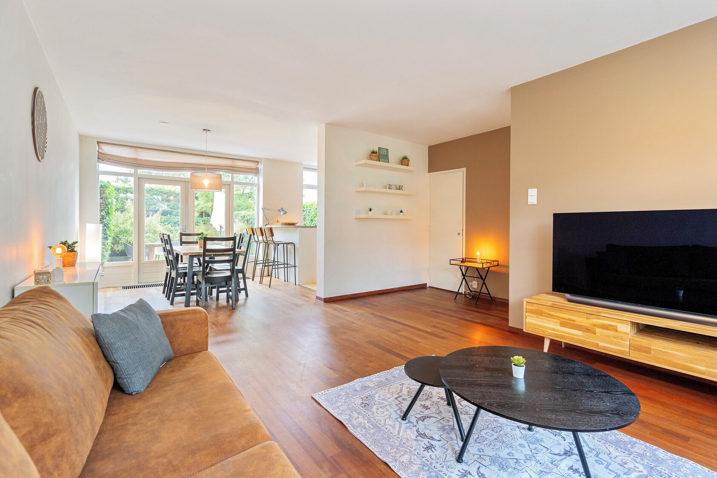 Een moderne woonkamer met een bank, een televisie, een eethoek met stoelen en een tafel, en een groot raam.