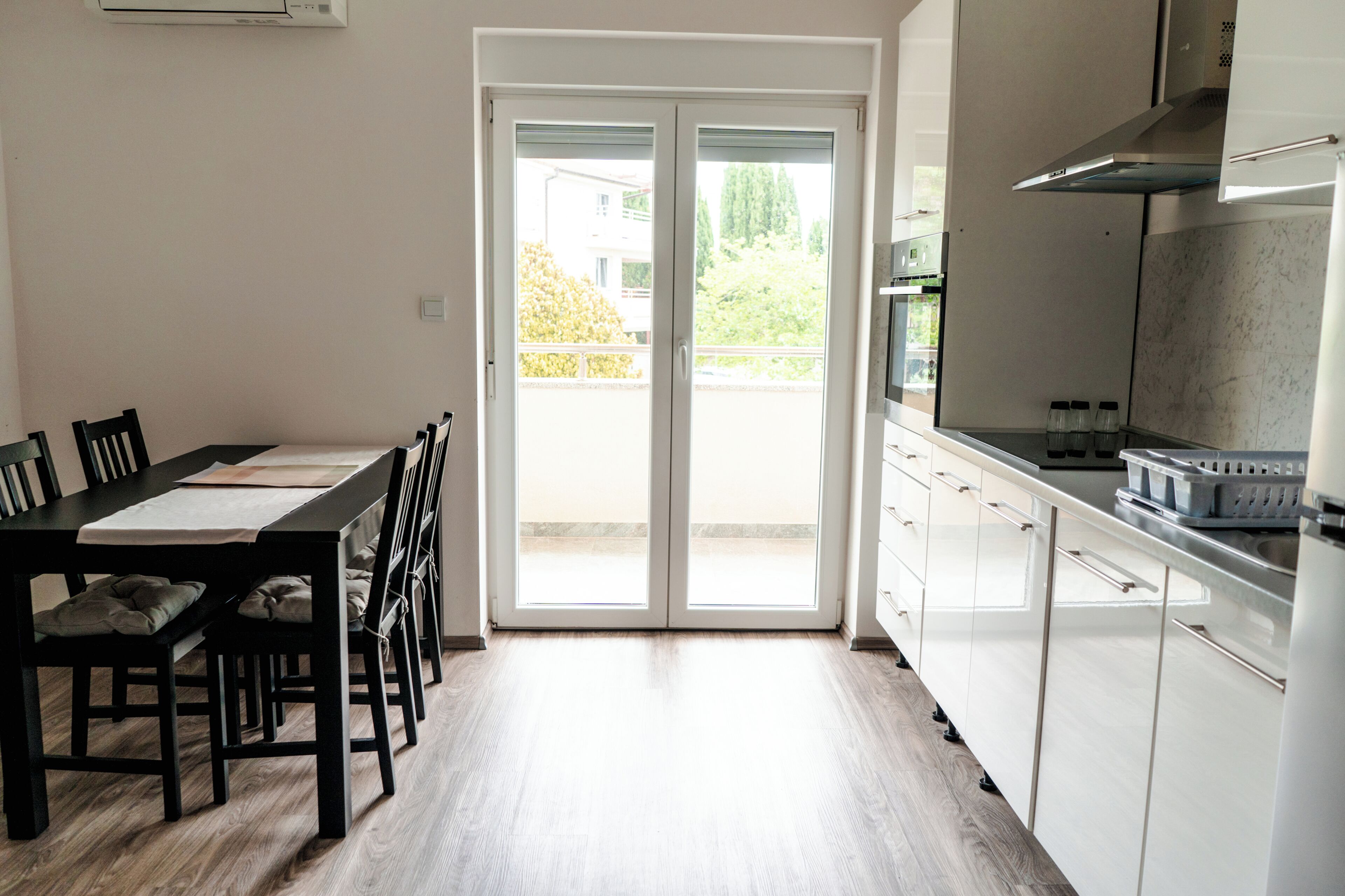 A modern kitchen with white cabinets, a dining area with a table and chairs, and a sliding glass door leading to a balcony.