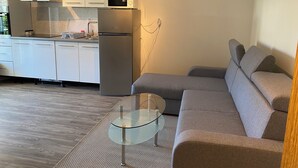 Eine moderne Küche mit weißen Schränken, einem Edelstahlkühlschrank und einer Mikrowelle darüber. Im Wohnbereich befindet sich ein graues Sofa und ein Glastisch.