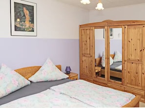 Un dormitorio con cama, armario de madera, silla de mimbre y un cuadro enmarcado en la pared.