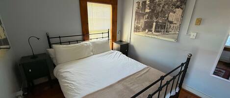 Une chambre à coucher comprenant un lit, une table de chevet, une lampe, une fenêtre avec des stores et un tableau accroché au mur.