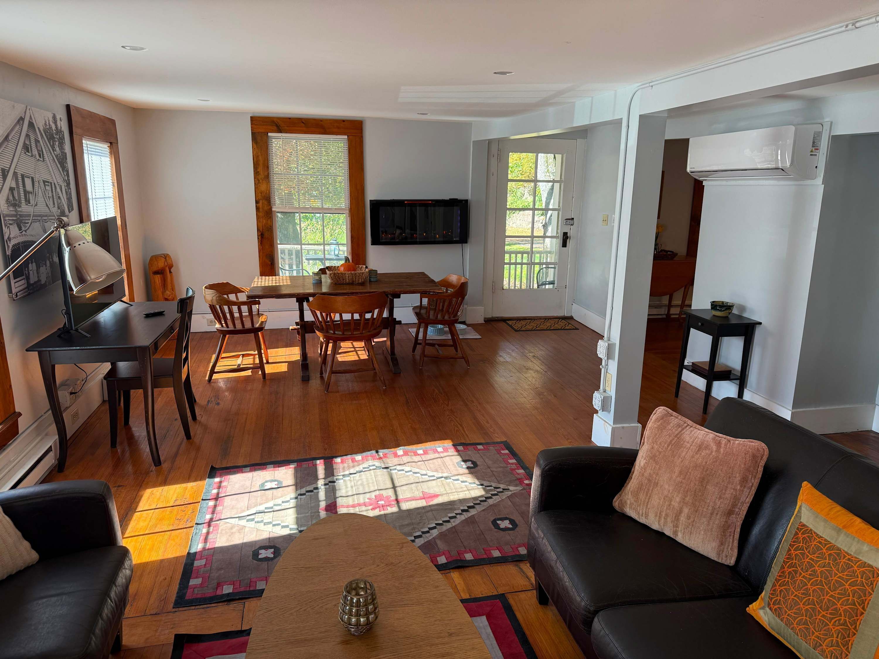 Sala de estar con sofá de cuero negro, mesa de centro de madera, comedor con mesa y sillas de madera, televisor montado en la pared y un ventanal con persianas.