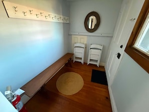 Quarto com um banco de madeira, duas cadeiras brancas, um tapete tecido, um espelho fixado na parede e um extintor de incêndio.