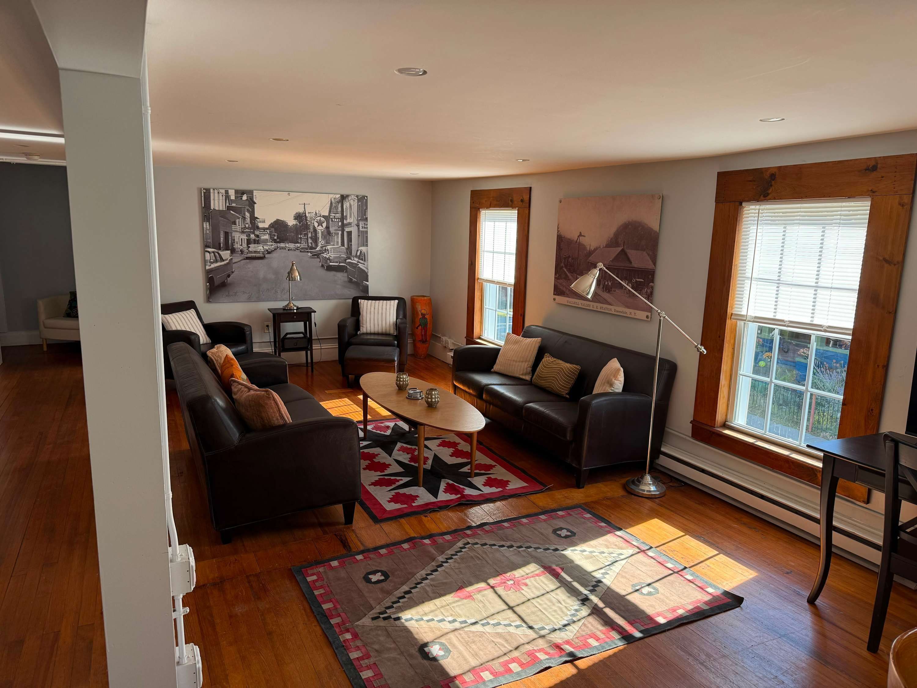 A living room with a sofa, armchair, coffee table, and two rugs. There are two windows with blinds, a lamp, and a painting on the wall.