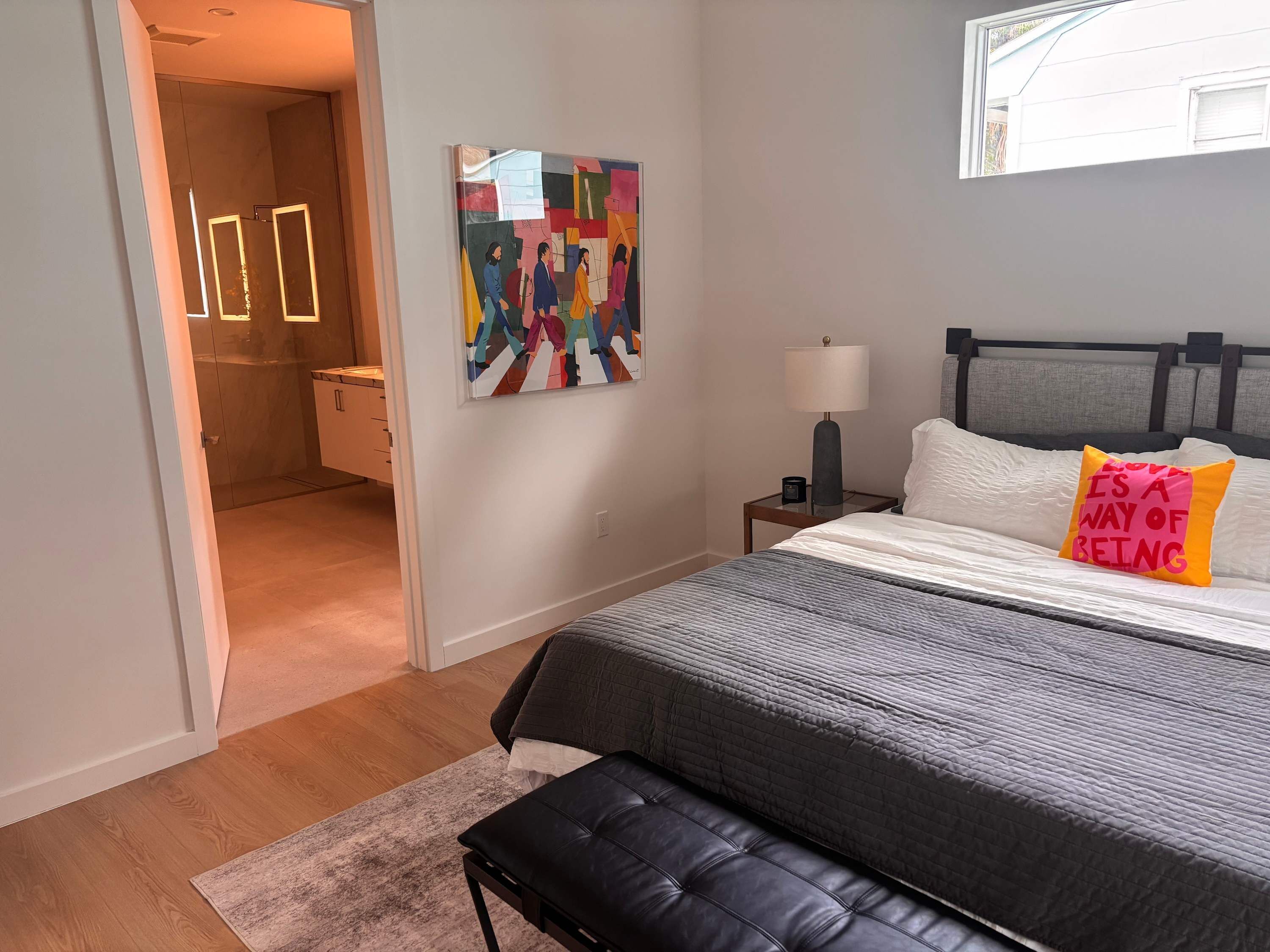 A bedroom with a bed, a bench, a bedside table with a lamp, a painting on the wall, and a bathroom visible through an open door.