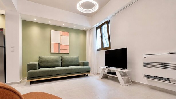 A modern living room with a sofa, a flat-screen TV on a stand, and a window. - Galleria Romvis – VΙ by ΜΡS (Athina)