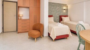 A compact room with a wardrobe, a single chair, two beds, a small table, and a wall-mounted light fixture. - Galleria Romvis – VΙ by ΜΡS (Athina)