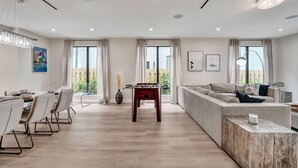 Una sala de estar moderna con un sofá, una mesa de comedor con sillas, una mesa de fútbol de mesa y ventanas amplias con cortinas.