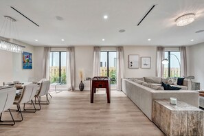 Una sala de estar moderna con un sofá, una mesa de comedor con sillas, una mesa de fútbol de mesa y ventanas amplias con cortinas.