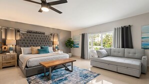 Une chambre moderne avec un lit, des tables de chevet, un ventilateur de plafond, un canapé gris et une grande fenêtre avec des rideaux.