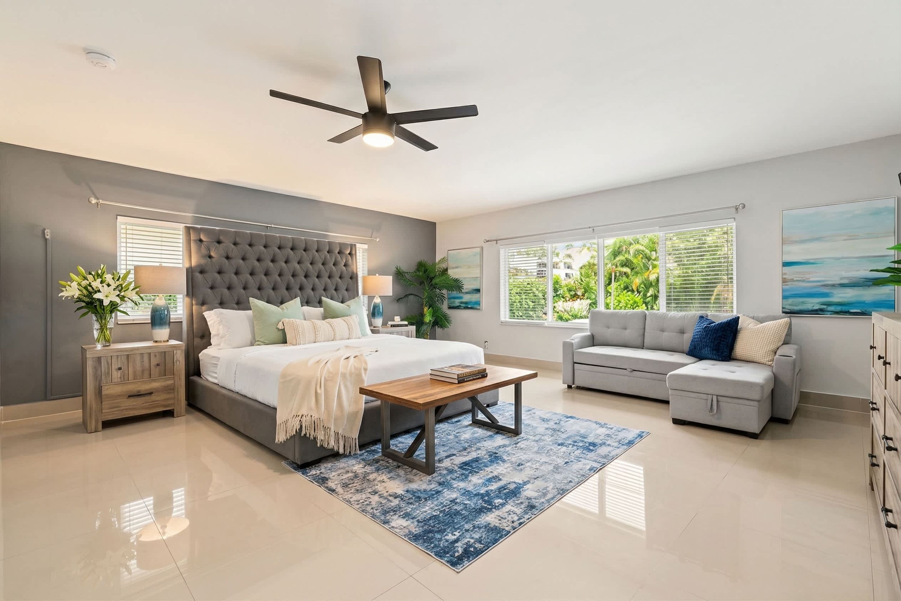 A bedroom with a bed, bedside tables, a sofa, a coffee table, a ceiling fan, and large windows.